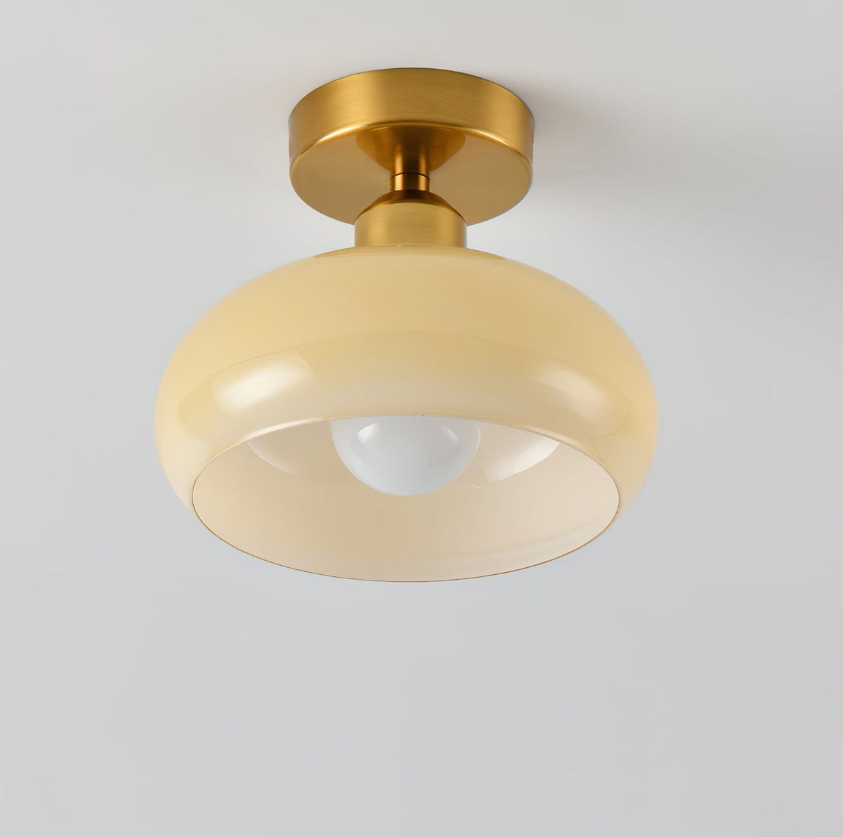 A Clérachic ceiling light with a pale beige glass dome and brass canopy, seen from slightly below against a light wall.