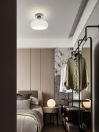 White glass Clérachic ceiling light above a dark-framed doorway, with a made bed and a green shirt on a rail beyond.