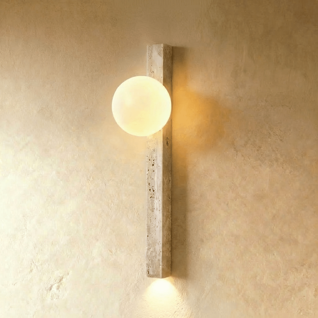 Clérachic slim beige travertine wall light with a lit opal globe, glowing warm with a soft downlight on a limewash wall.