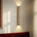 Clérachic travertine column wall light on a white plastered wall above a red velvet banquette, with sunlight from a curtained window.