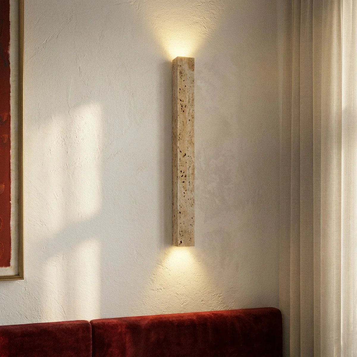 Clérachic travertine column wall light on a white plastered wall above a red velvet banquette, with sunlight from a curtained window.