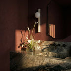 Clérachic travertine wall light with an opal globe in a dark red bathroom, beside a mirror, stone basin and white calla lilies.