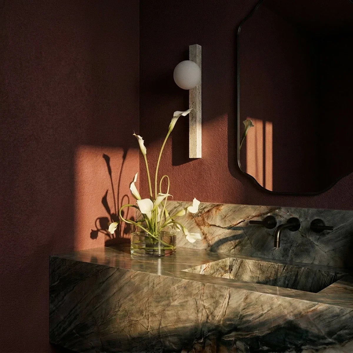 Clérachic travertine wall light with an opal globe in a dark red bathroom, beside a mirror, stone basin and white calla lilies.