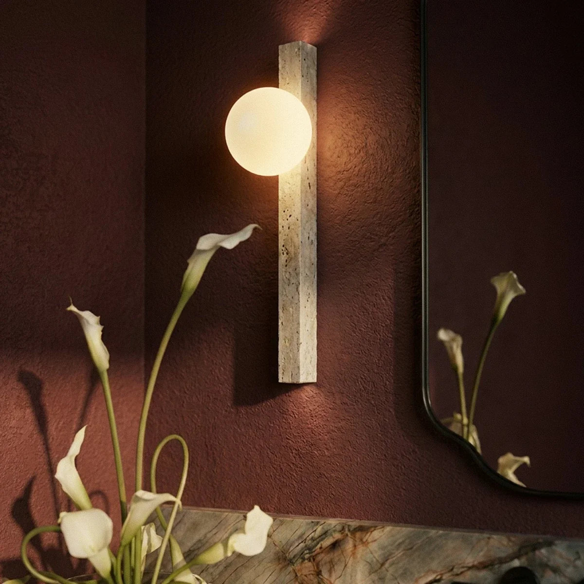 Clérachic close-up of a glowing opal globe on a travertine column wall light, set against a textured red wall and calla lilies.