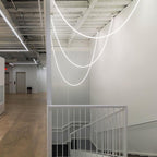 Several Clérachic glowing LED rope pendants swooping over a white stairwell, beside a white railing in a concrete-floored space.
