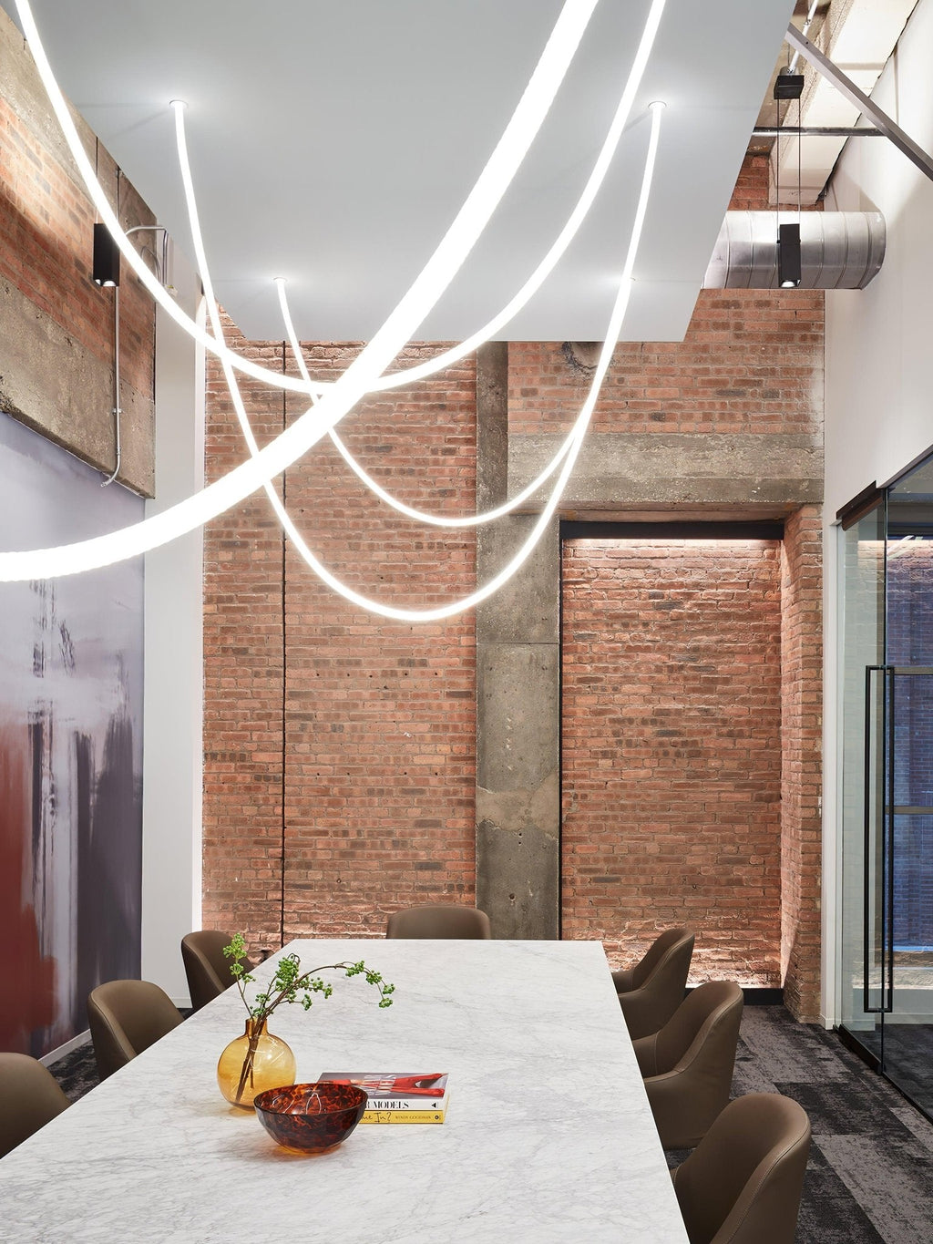 Bright Clérachic LED rope pendants swooping through a brick-walled room above a marble table with a vase and a bowl.