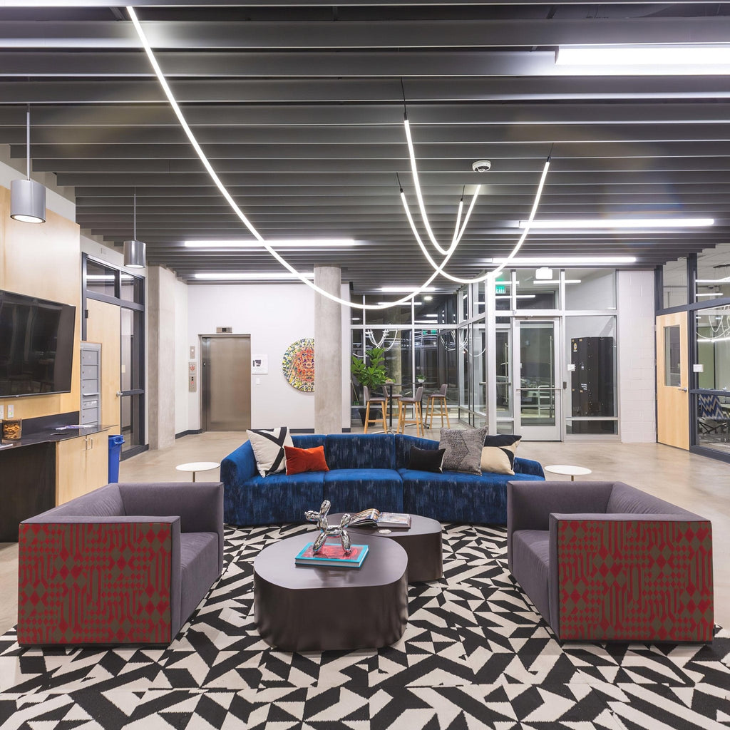 A Clérachic LED rope pendant arcing across a dark industrial ceiling above a blue sofa and two grey armchairs on a patterned rug.