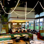 Looping Clérachic LED rope pendants arching high above a restaurant bar with green banquettes and gold globe lights.