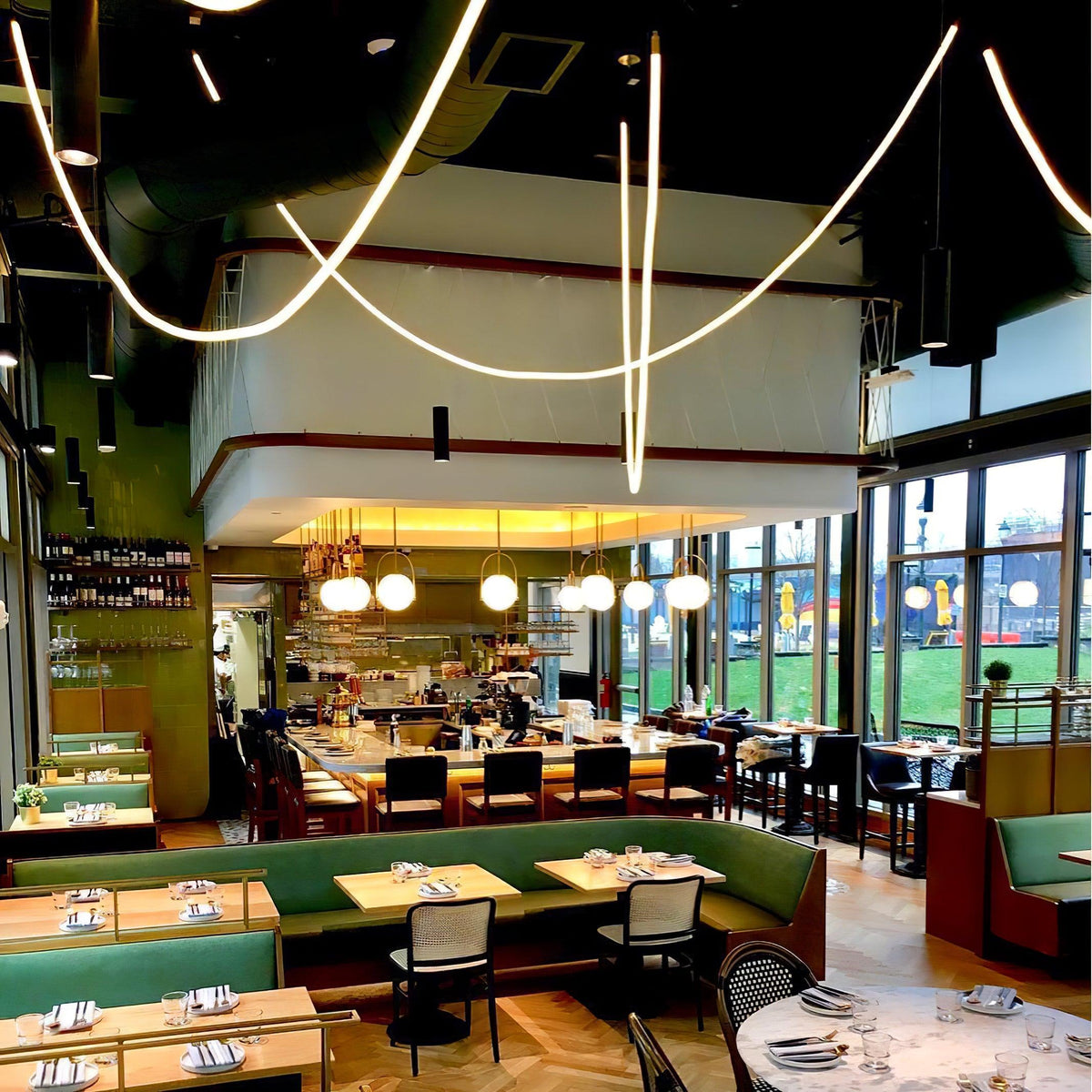 Looping Clérachic LED rope pendants arching high above a restaurant bar with green banquettes and gold globe lights.