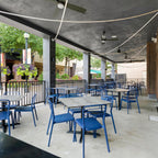 A Clérachic rope pendant arcing across the ceiling of a covered terrace above blue chairs, with fans and a leafy street behind.