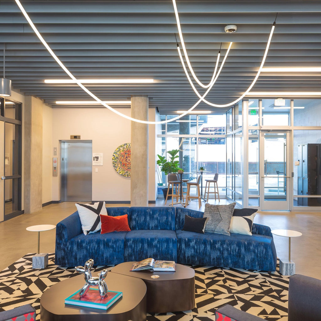 Clérachic LED rope pendants curving under a slatted ceiling above a blue sofa and a patterned rug, near a glass entrance.