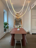 Glowing Clérachic LED rope pendants swooping above a dining table with a terracotta top and white chairs, in front of a framed figure.