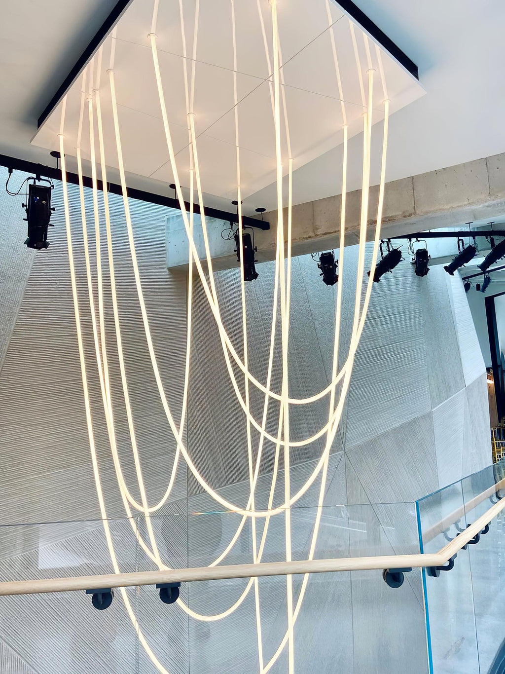 Many long Clérachic LED rope pendants dropping through a white square opening above a stairwell with a glass balustrade.