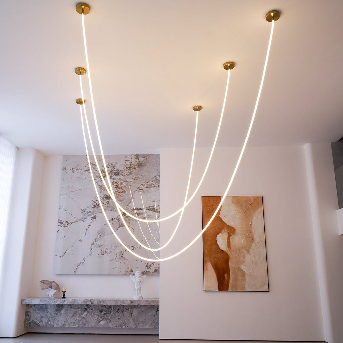 Several Clérachic LED rope pendants with gold-toned plates at different heights, curving in front of a marble wall and a rust-coloured painting.