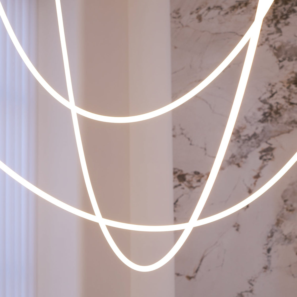 Close view of the soft, even glow along the tubes of Clérachic LED rope pendants, with a marble wall and a curtain behind.