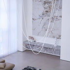 Unlit Clérachic rope pendants as white lines in front of a marble wall, sheer curtains and a grey marble shelf.