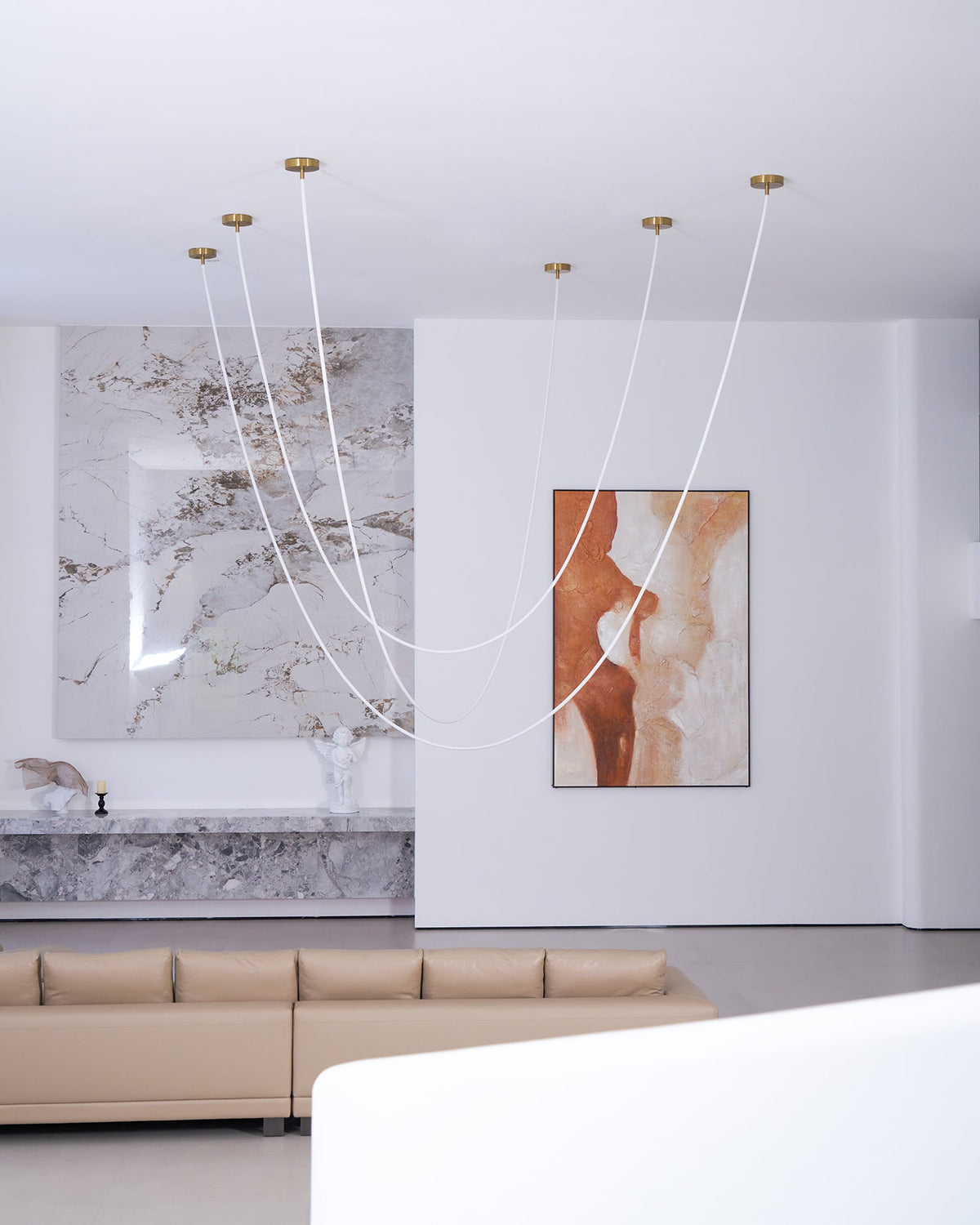 Unlit Clérachic rope pendants hanging from gold-toned plates before a marble feature wall, a painting and a taupe sofa.