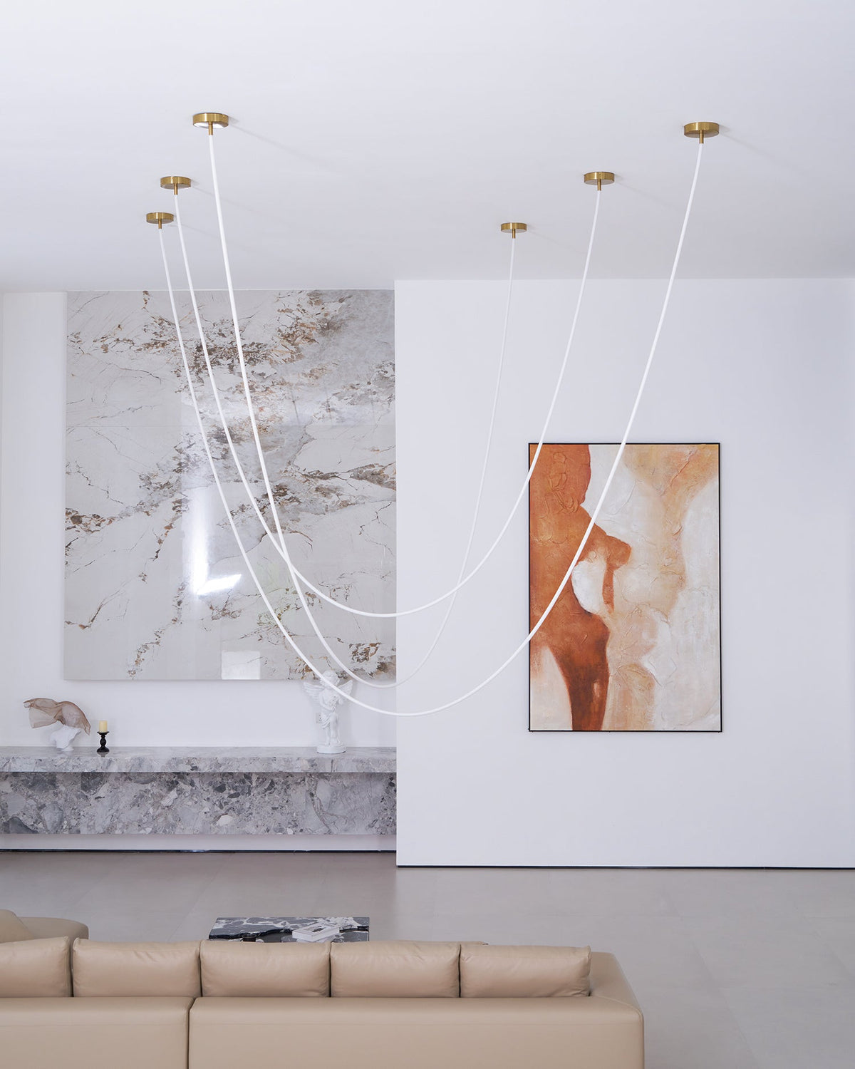 Pale, unlit Clérachic rope pendants in loops in a high white living room, with a taupe sofa below a marble feature wall.