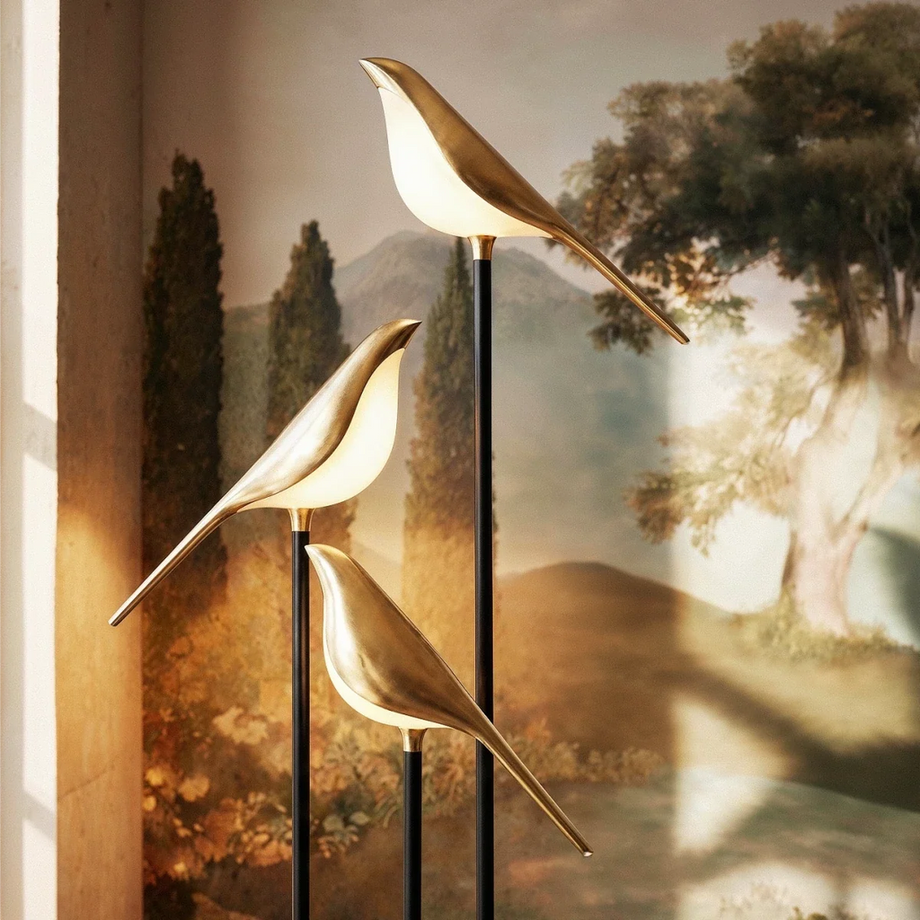 Close view of three Clérachic gold bird shades glowing warm white on black stems against a landscape mural