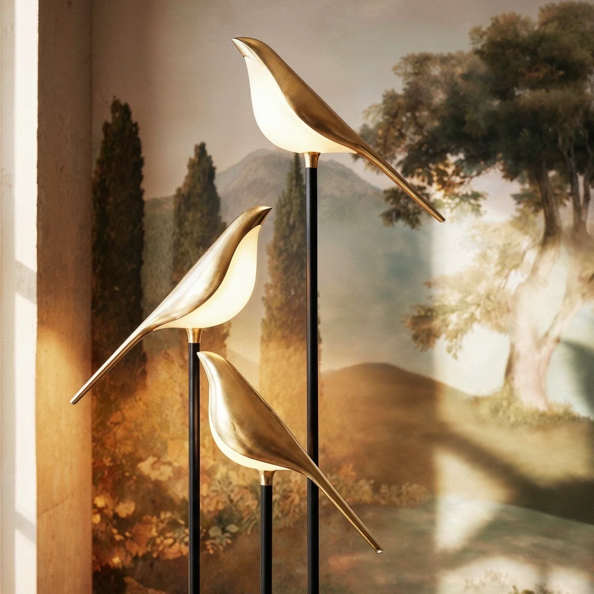 Close view of three Clérachic gold bird shades glowing warm white on black stems against a landscape mural