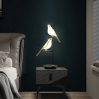 Clérachic two-head bird table lamp lit on a dark bedside cabinet against a deep green wall, beside a leather headboard.