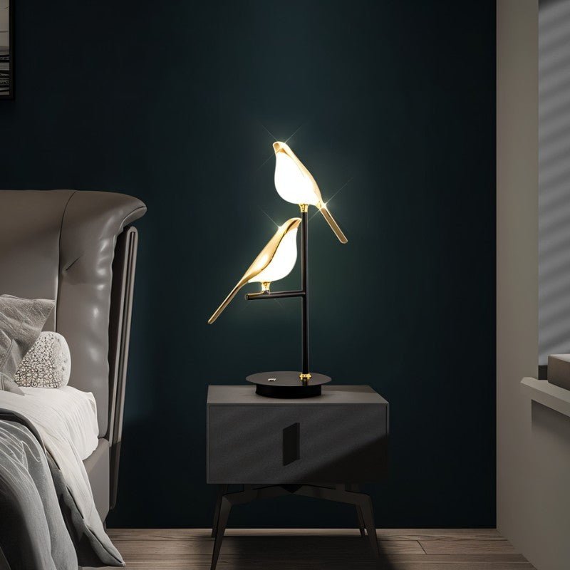 Clérachic two-head bird table lamp lit on a dark bedside cabinet against a deep green wall, beside a leather headboard.