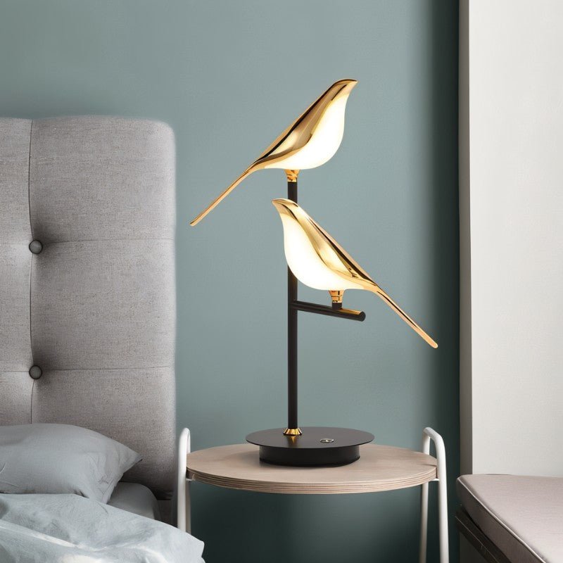 Clérachic two-head bird table lamp with gold birds on a round wooden bedside table, against a muted blue-green wall.