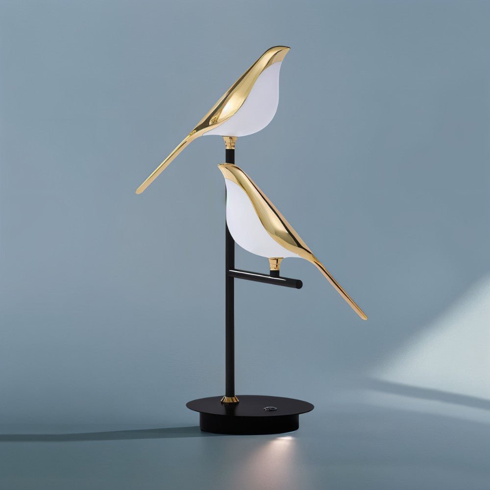 Clérachic two-head bird table lamp with polished gold birds and white acrylic shades on a black round base, on soft blue-grey.