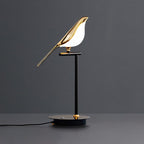 Clérachic one-head bird table lamp with a lit gold bird on a black stem, base cable trailing across a dark grey surface.