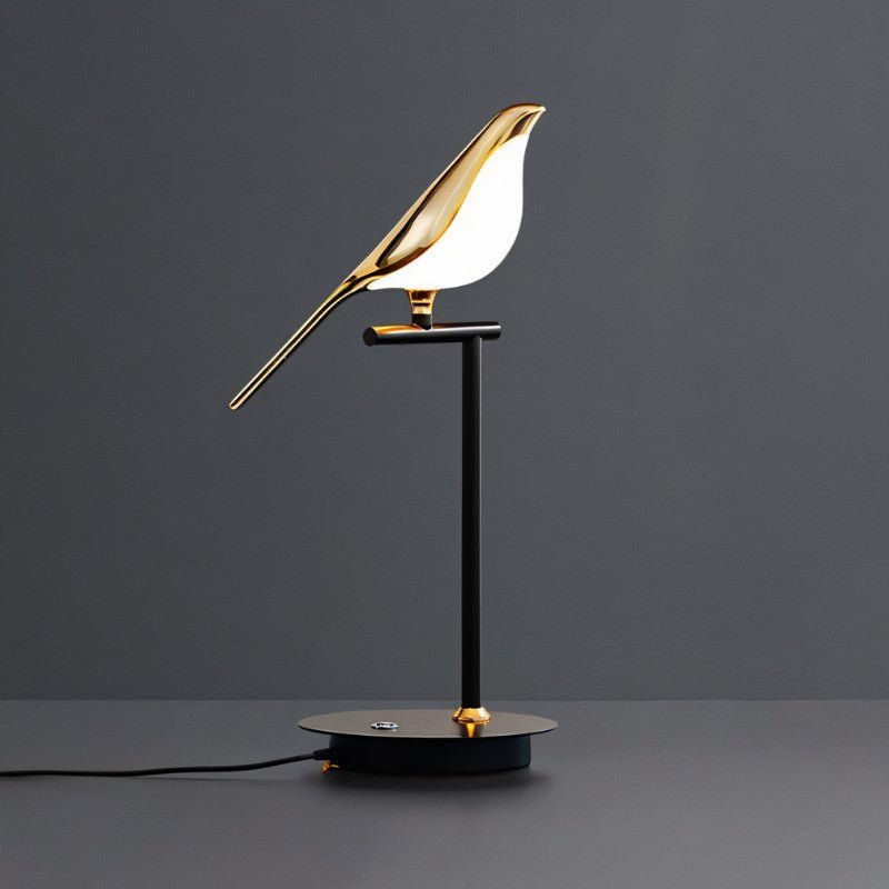 Clérachic one-head bird table lamp with a lit gold bird on a black stem, base cable trailing across a dark grey surface.