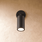 Clérachic black aluminium wall spotlight on a round backplate, its cylinder angled down with the LED glowing warm on plaster.