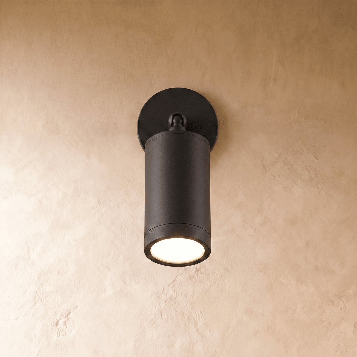 Clérachic black aluminium wall spotlight on a round backplate, its cylinder angled down with the LED glowing warm on plaster.