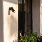 Clérachic black wall spotlight tilted downward and glowing warm on a sunlit white wall next to a black-framed window.