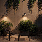Two Clérachic black wall spotlights lit warm, each casting a pool of light down a stone wall above chairs and a leafy plant.