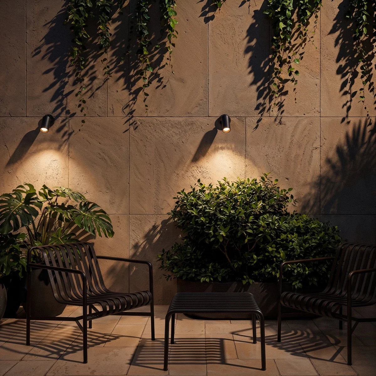Two Clérachic black wall spotlights lit warm, each casting a pool of light down a stone wall above chairs and a leafy plant.