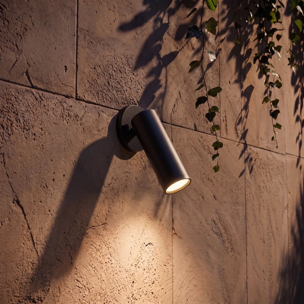 Close-up of a Clérachic black cylinder wall spotlight angled down, its warm LED lighting the textured stone wall below.