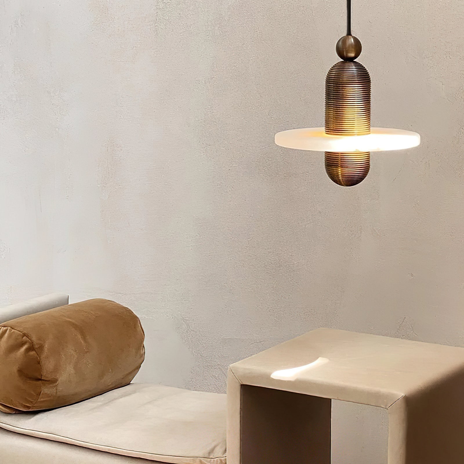 Clérachic gold single-disc pendant glowing above a cream daybed and tan side table on a plaster wall.