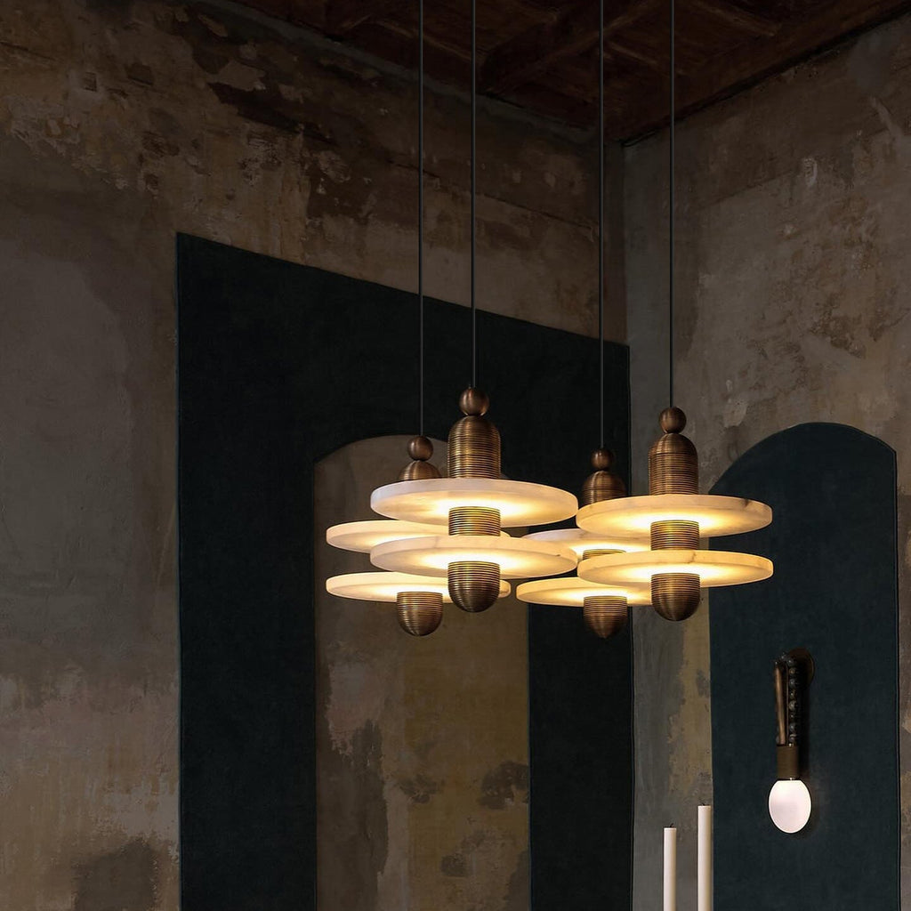 Clérachic gold double-disc pendants cluster four-high before a teal arched niche on a distressed plaster wall.