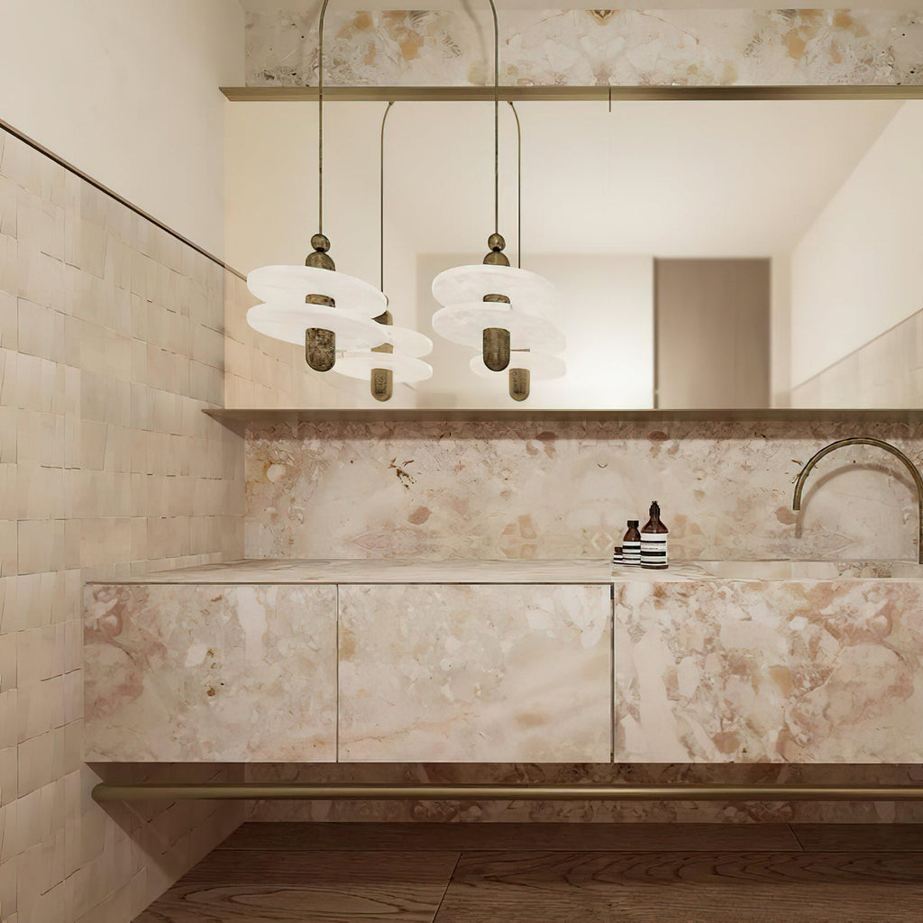 Clérachic gold double-disc pendants hang above a pink-veined marble bathroom vanity and arched gold faucet.