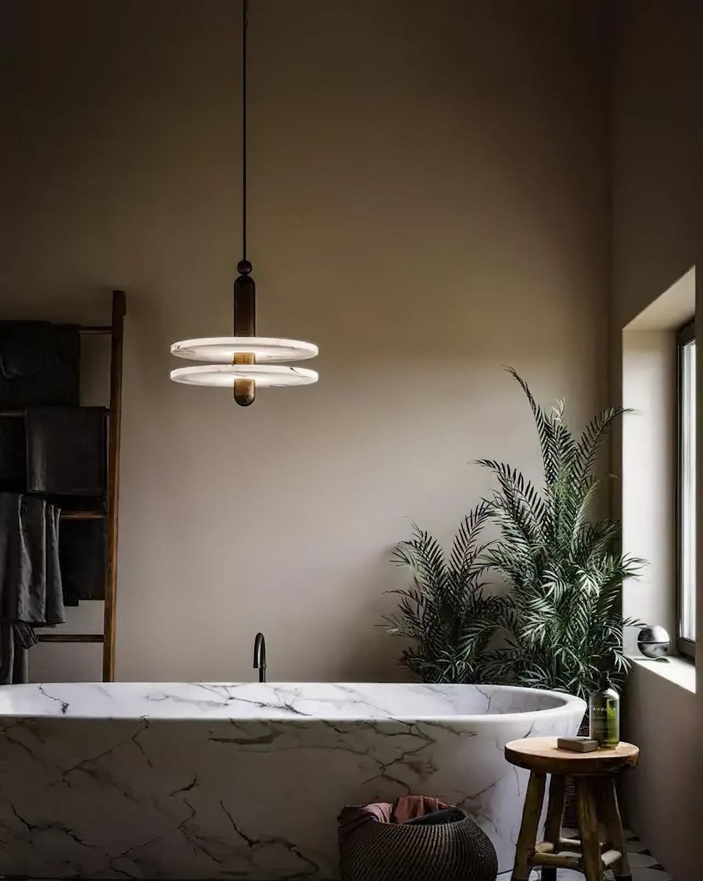 Clérachic gold double-disc pendant hangs above a veined marble bathtub beside a wooden stool and palm.
