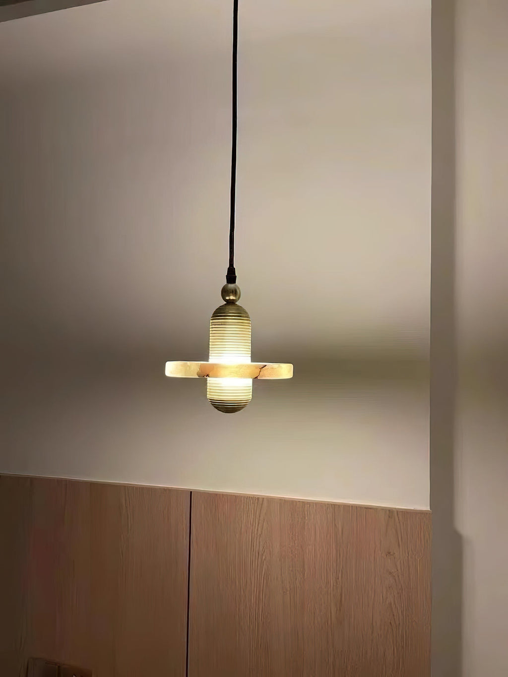 Clérachic gold single-disc pendant glowing beside a cream wall and light wood panel, used as bedroom light.