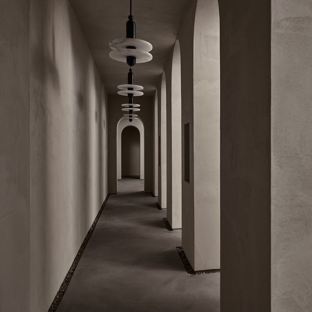 Clérachic pendants with stacked alabaster discs on black stems line a taupe arched hallway over pebbles.