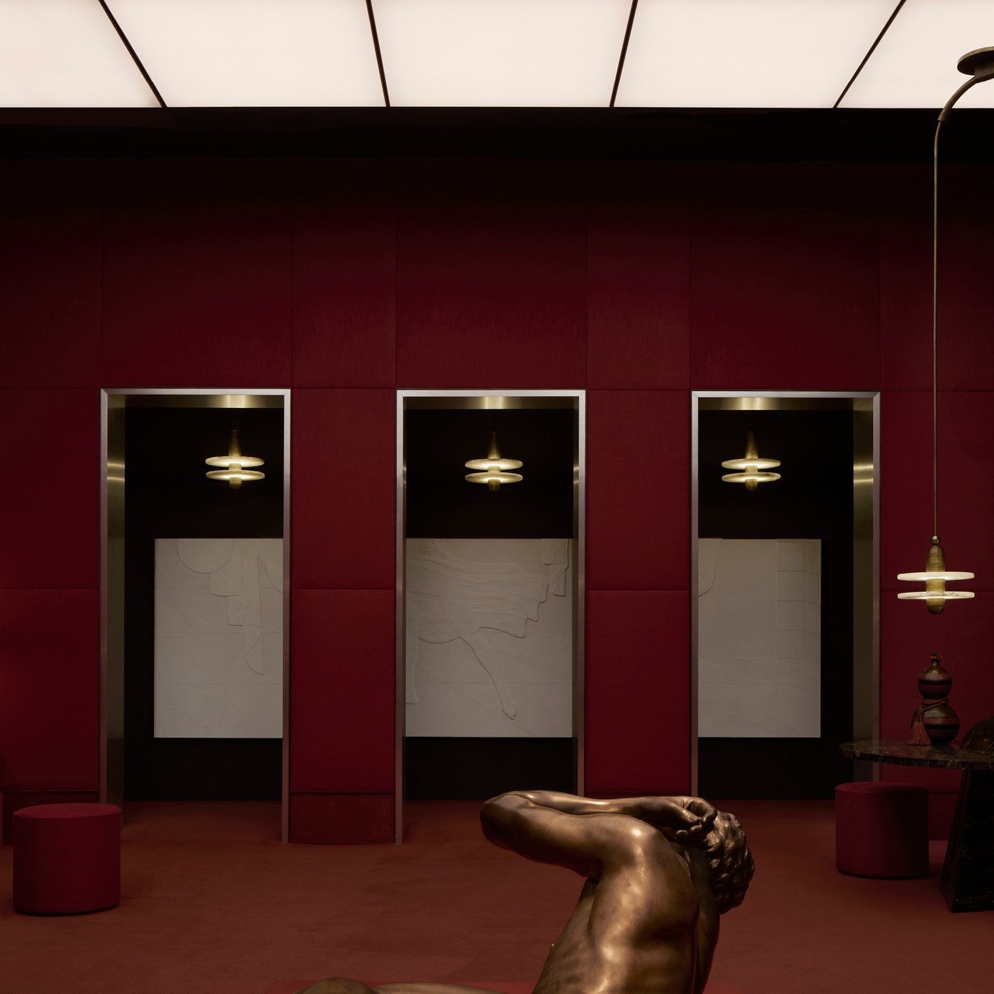 Clérachic gold double-disc pendants hang in red-paneled alcoves above a bronze reclining figure.