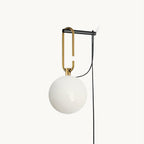 A Clérachic wall lamp with a frosted white glass globe hanging from a black and gold hook frame, its black cord looping down, on white.