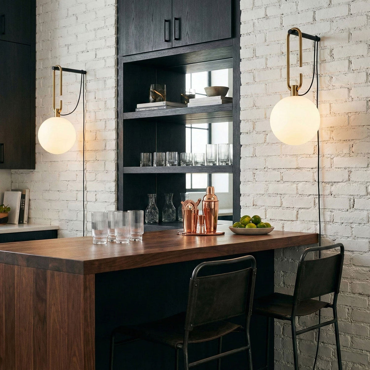 Two lit Clérachic globe wall lamps hang on a white brick wall above a walnut bar counter with glasses and dark metal stools.