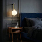 A lit Clérachic glass globe lamp on a charcoal panelled wall beside a navy bed and a walnut bedside table with a decanter.
