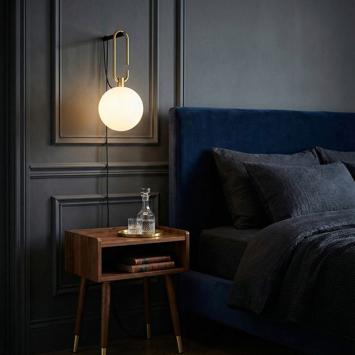 A lit Clérachic glass globe lamp on a charcoal panelled wall beside a navy bed and a walnut bedside table with a decanter.
