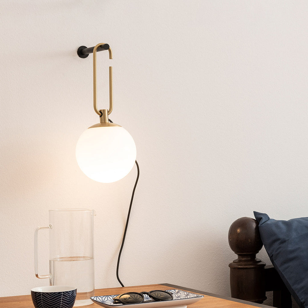 A Clérachic globe wall lamp lit white above a wooden side table with a glass jug, a patterned bowl and a pair of sunglasses.