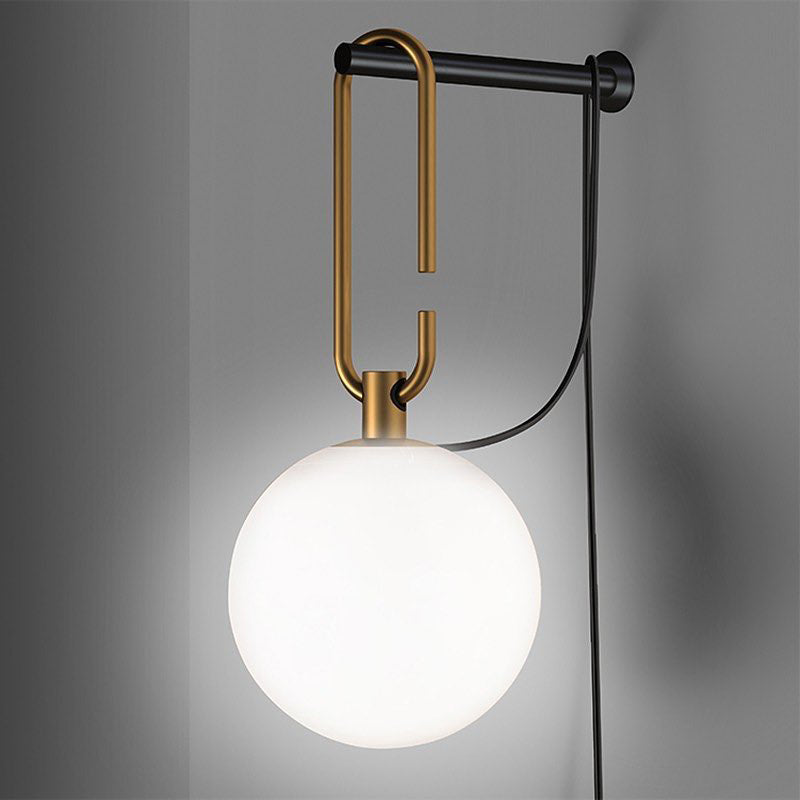 A bright white globe hangs from a gold loop on a black wall bar, with the black cord curving away, on a grey wall, from Clérachic.