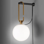 A bright white globe hangs from a gold loop on a black wall bar, with the black cord curving away, on a grey wall, from Clérachic.